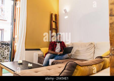 Black man in casual clothes using laptop on the sofa at home. He is surfing the net visiting some websites. Stock Photo