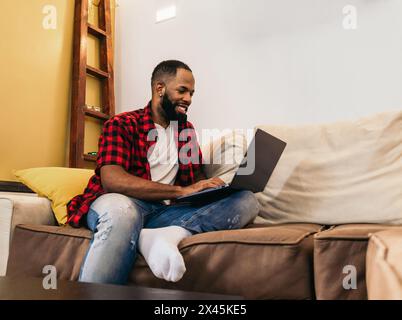 Smiling black man using laptop sitting on the sofa at home. He looks relaxed and happy. Stock Photo