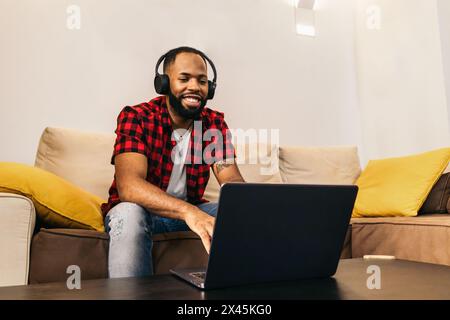 Black man in casual clothes and headphones sitting on the sofa in front of a laptop. The image conveys the concept of watching movies and videos on th Stock Photo