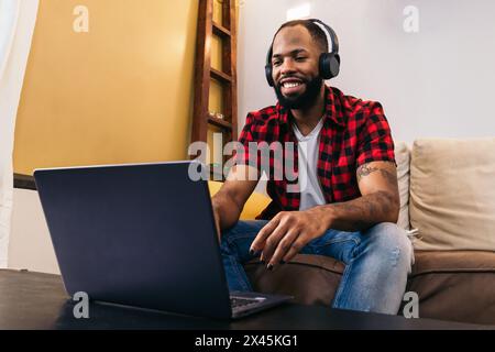 Black man with painted nails in casual clothes and headphones sitting on the sofa in front of a laptop. The image conveys the concept of watching movi Stock Photo
