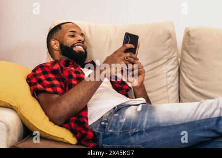 Black man with beard and painted nails using smartphone on the sofa at home. He wears casual clothes and is lying on the sofa relaxing watching videos Stock Photo