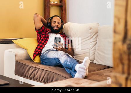 Black man with beard and painted nails using smartphone and headphones on the sofa at home. He wears casual clothes and is lying on the sofa relaxing Stock Photo