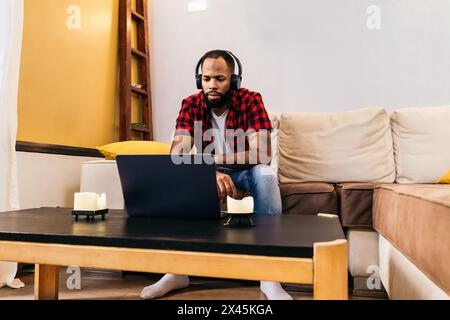 Black man with painted nails in casual clothes and headphones sitting on the sofa in front of a laptop. The image conveys the concept of watching movi Stock Photo