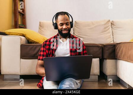 Black man in casual clothes using laptop at home. He is sitting on the floor leaning on the sofa in the living room while he listens to music with hea Stock Photo