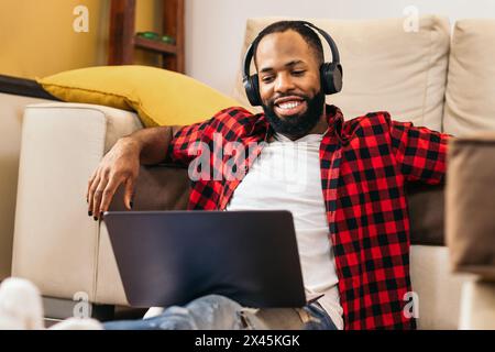Black man in casual clothes using laptop at home. He is sitting on the floor leaning on the sofa in the living room while he listens to music with hea Stock Photo
