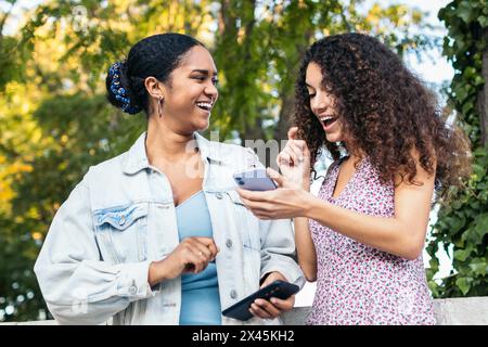 Two Latin American friends walking down the street, having fun, and laughing while using a smartphone. Stock Photo