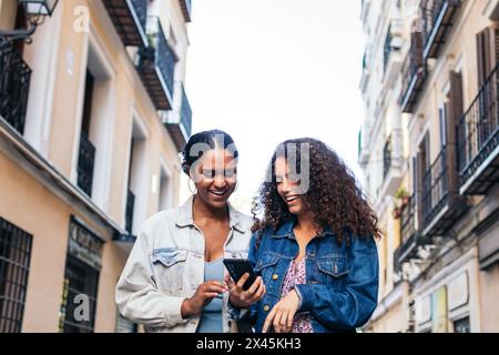 Two Latin American friends walking down the street, having fun, and laughing while using a smartphone. Stock Photo