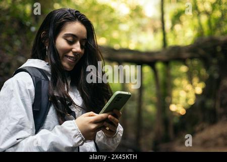 Young woman in raincoat and backpack using smartphone while walking in the forest Stock Photo