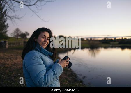 Young photographer woman making sunset photos of a nice lake in nature. She looks at camera smiling while reviews the pictures she has taken Stock Photo