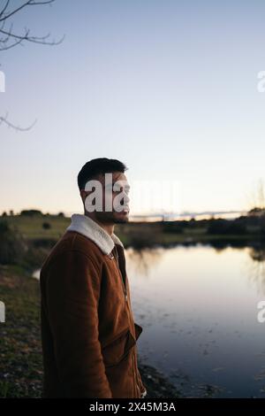 Attractive young man with a beard watching the sunset at a lake in the countryside. He looks calm enjoying the beautiful scenery in nature Stock Photo