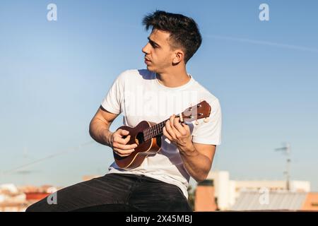 young man sitting playing ukelele on a rooftop Stock Photo