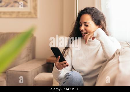 Young woman sitting on a comfortable sofa in her home, using her mobile phone with a relaxed expression on her face. Stock Photo