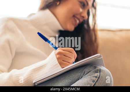 Young woman sitting in her home, studying for university with her laptop and a notebook. The student is taking notes while reviewing information onlin Stock Photo