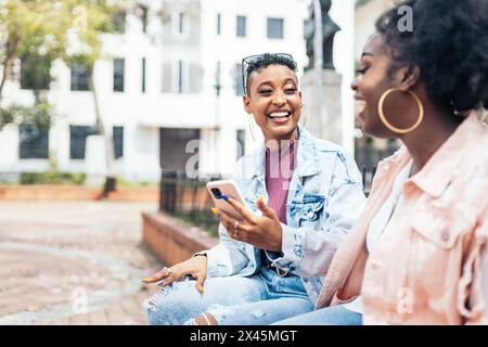 Two black women friends using smartphone and talking sitting on a bench laughing and having fun in the city Stock Photo