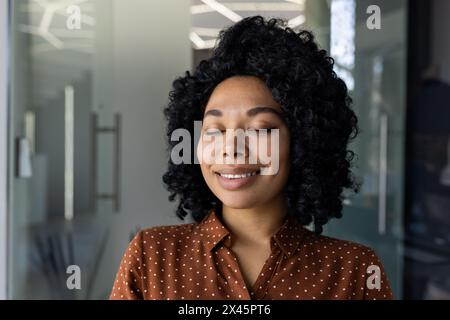 A young African-American woman with curly hair, dressed in a polka dot shirt, closes her eyes and smiles, enjoying a serene moment in a sunlit modern office environment. Stock Photo