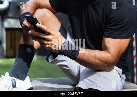 Cropped unrecognizable muscular sportsman in sportswear using cellphone while resting after intense workout in gym Stock Photo