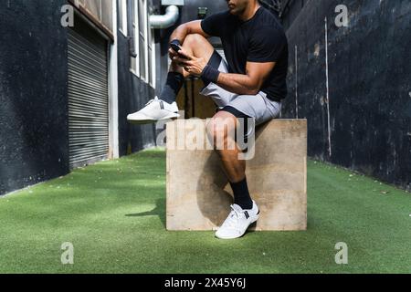 Cropped unrecognizable muscular sportsman in sportswear using cellphone while resting after intense workout in gym Stock Photo
