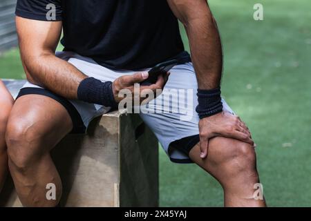 Cropped unrecognizable muscular sportsman in sportswear using cellphone while resting after intense workout in gym Stock Photo
