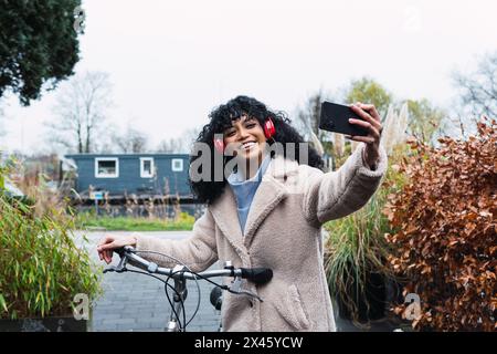 Cheerful young African American female with curly dark hair in warm coat walking with bicycle while taking selfie and listening to music in headphones Stock Photo