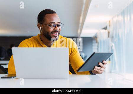 Happy black male sitting at table with laptop and listening for music in earphones from tablet in coworking space Stock Photo