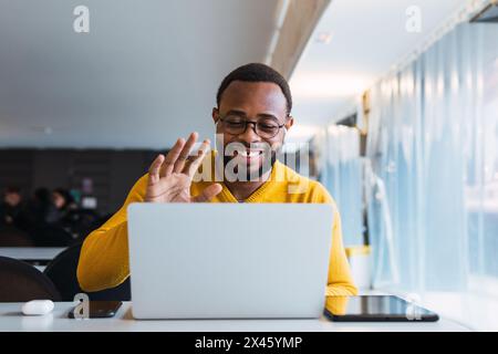 Cheerful African American bearded male entrepreneur in sweater and eyeglasses having video call on netbook with earphones while working in office Stock Photo