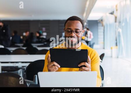 Happy black male sitting at table with laptop and listening for music in earphones from tablet in coworking space Stock Photo