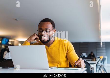 Positive bearded African American male entrepreneur taking notes on tablet using stylus while working on netbook in office and looking at screen touch Stock Photo