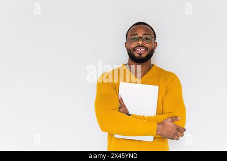 Positive bearded African American male in yellow sweater and eyeglasses holding notebook while looking at camera and standing against white background Stock Photo