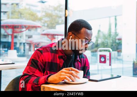 Side view of concentrated African American man in checkered shirt watching video on tablet with earphones while drinking hot beverage in cafe sitting Stock Photo