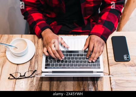 From above cropped unrecognizable African American bearded male freelancer browsing netbook with earphones while working remotely in cafe and looking Stock Photo