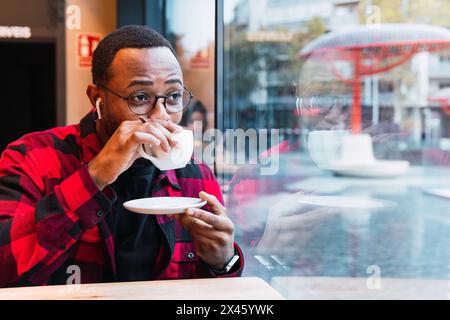 Thoughtful bearded African American male in checkered shirt with earphones drinking hot beverage from cup while resting in cafe Stock Photo