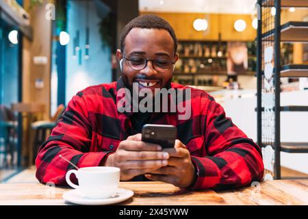 Smiling African American bearded male in red checkered shirt messaging on mobile phone wit earphones while drinking coffee in cafe and looking at scre Stock Photo