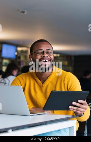 Happy black male sitting at table with laptop and listening for music in earphones from tablet in coworking space Stock Photo