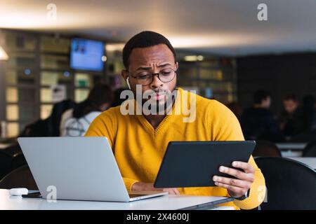 Thoughtful black male sitting at table with laptop and listening for music in earphones from tablet in coworking space Stock Photo