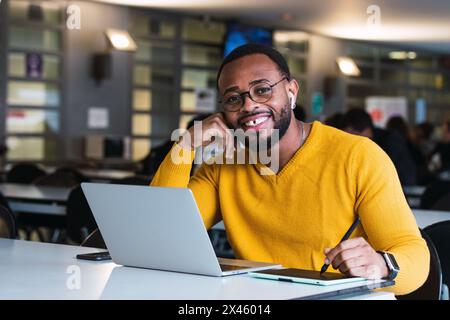 Positive bearded African American male entrepreneur working on graphic on tablet using stylus while on netbook in office and looking at camera Stock Photo