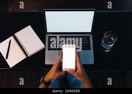 From above of crop unrecognizable male worker text messaging on cellphone with empty screen while sitting at table with netbook Stock Photo