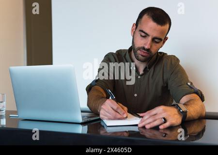 Focused male taking notes in notepad while sitting at table with modern netbook in light room during work time Stock Photo