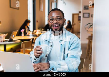 Smiling African American male with cup of coffee looking away while surfing netbook at table in modern cafe during break Stock Photo