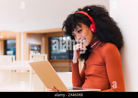 Black female student in headphones sitting and browsing laptop in university library Stock Photo