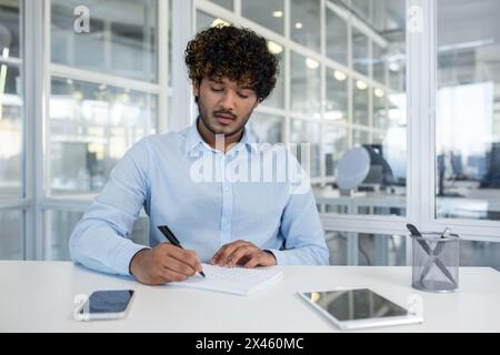 Young professional male in a modern office setting, intently writing in a notebook with a smartphone and computer nearby. Represents productivity and workplace concentration. Stock Photo