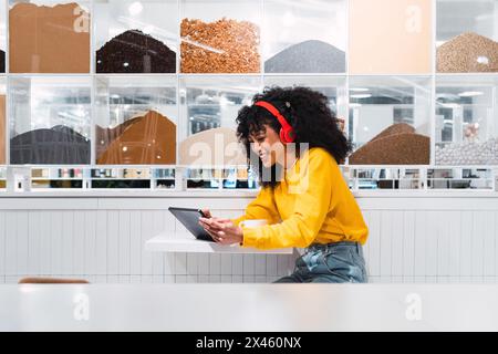 Black lady with curly hair smiling while sitting at table listening to music from tablet Stock Photo
