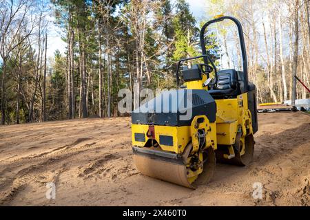 Closeup of yellow compactor roller, a machined used for compaction of soil, asphalt surface, gravel and sand. Viewed on an empty, tree lined building Stock Photo