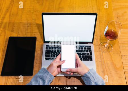 From above anonymous female freelancer browsing smartphone with blank screen over laptop near tablet and red wine while sitting at table in restaurant Stock Photo