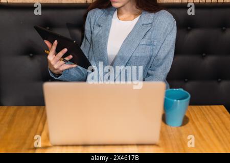 Cropped unrecognizable young female entrepreneur browsing data on tablet while sitting at table with laptop and a cup with hot beverage during remote Stock Photo