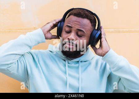 Happy adult bearded African American male in blue hoodie enjoying music with wireless headphones against yellow background Stock Photo