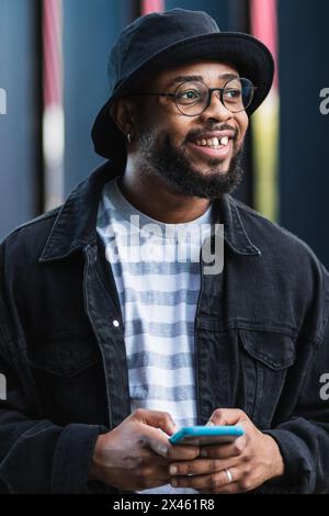 Cheerful adult bearded African American male in casual outfit and hat standing against wall and messaging on smartphone Stock Photo