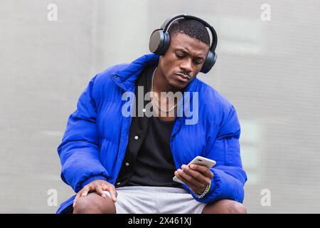 Calm ethnic male wearing stylish hipster outfit sitting on stone border on street in headphones and using mobile phone while looking away Stock Photo