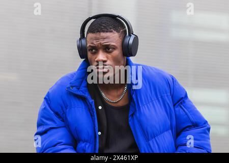 Calm ethnic male wearing stylish hipster outfit sitting on stone border on street in headphones and enjoying songs while looking away Stock Photo