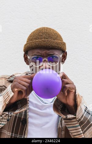 Young black male in stylish hat and glasses looking at camera and blowing violet balloon against gray wall Stock Photo