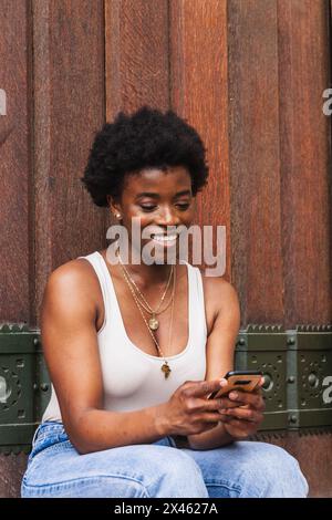 Cheerful curly haired African American female browsing social media in mobile phone and smiling brightly Stock Photo